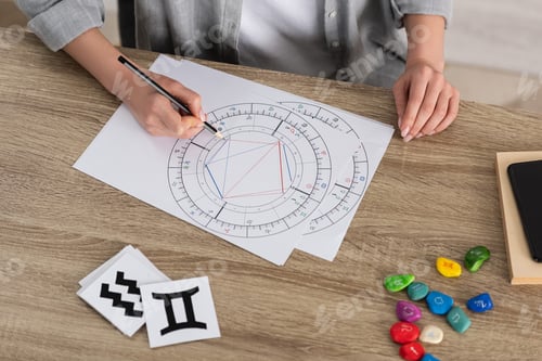 Preview: Cropped view of woman drawing natal chart by stones and papers with zodiac signs on table