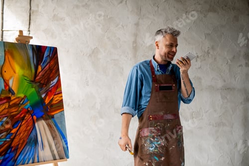 Preview: Cheerful middle aged painter in workwear recording voice message on smartphone