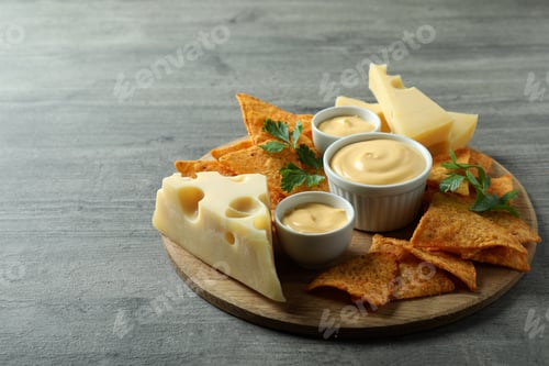 Preview: Board with cheese sauce, chips, cheese and parsley on gray textured table