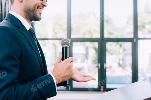 Preview: cropped image of smiling speaker gesturing and talking into microphone at podium tribune during