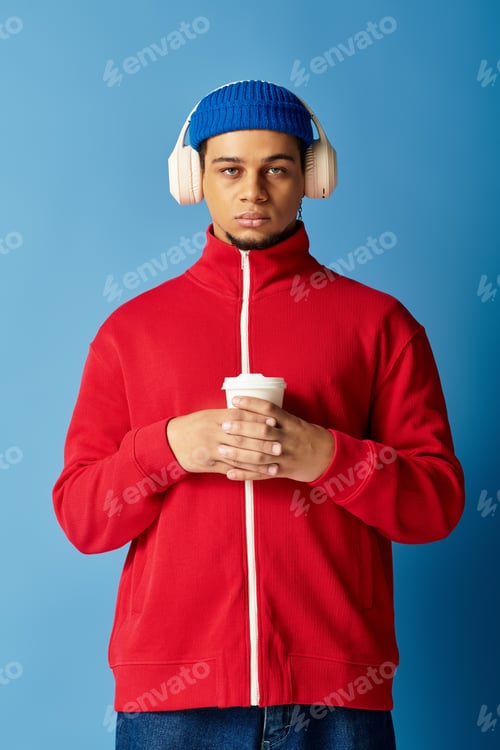 Preview: Stylish young man with beanie hat enjoying a drink against a vibrant blue background
