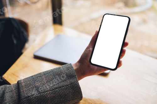 Preview: Mockup image of a woman holding mobile phone with blank white desktop screen with laptop on table