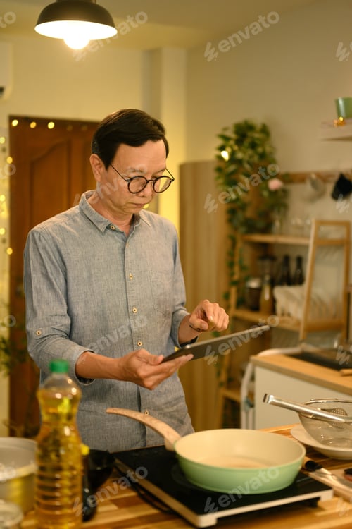 Preview: Portrait of senior man standing in warm kitchen, reading online recipe on digital tablet