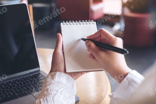Preview: Closeup image of hands holding and writing down on a blank notebook with computer laptop on table