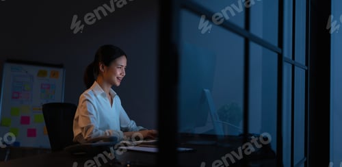 Preview: Smiling Asian business woman sitting the table and working on computer in office at night.