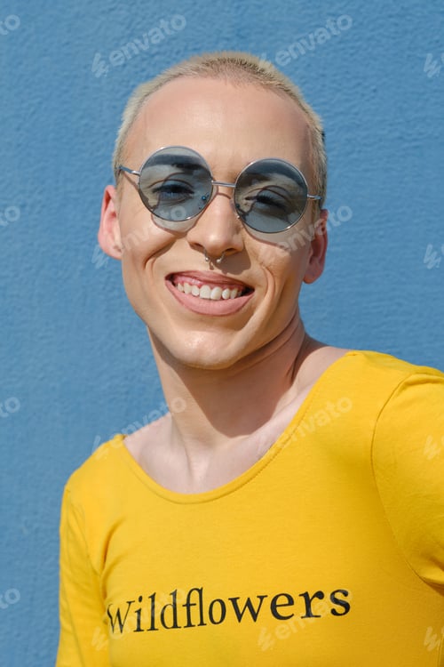 Preview: Close up view of a young non-binary person wearing sunglasses and smiling while posing outdoors.
