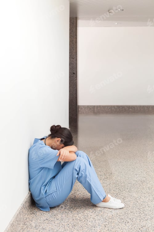 Preview: Asian woman in blue scrubs sitting on floor of hospital corridor leaning against wall, copy space