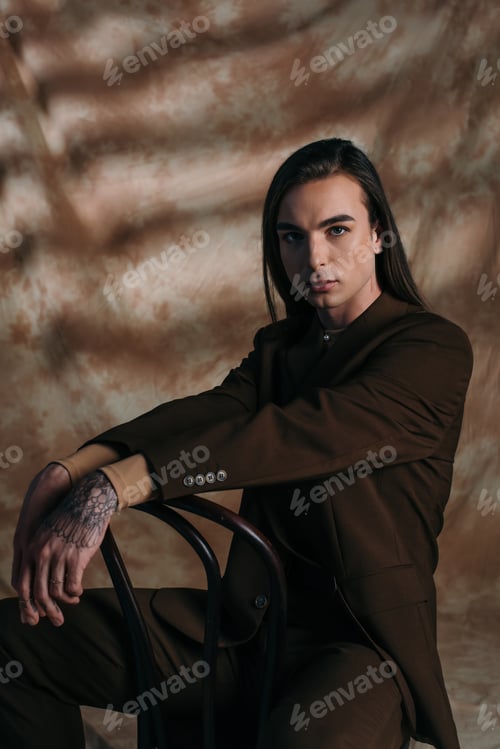 Preview: Portrait of trendy young nonbinary person sitting on chair on abstract brown background