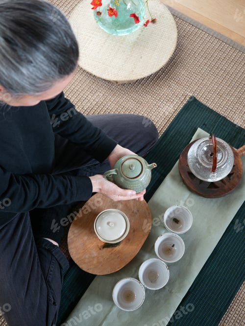 Preview: middleaged woman with gray hair sitting on the ground holding a small teapot preparing tea