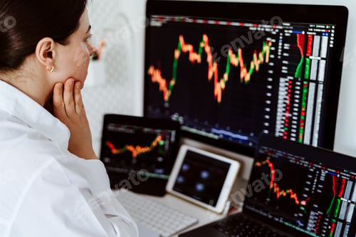 Preview: Young female cryptocurrency trader sitting at desk looking at multiple screens monitoring stock