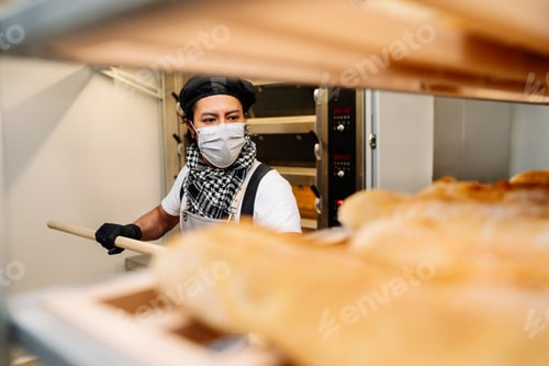 Preview: Latino baker shelving freshly baked bread with a protective face mask