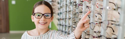 Preview: joyful girl looking at camera while choosing eyeglasses in optics store, banner