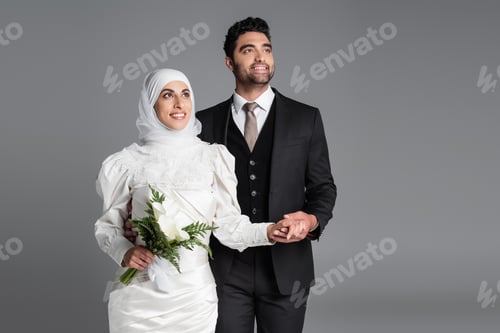 Preview: happy groom in suit holding hand of dreamy muslim bride with wedding bouquet of calla lily flowers