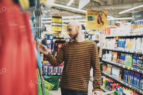 Preview: Adult bald bearded man looking by price in hardware department of supermarket