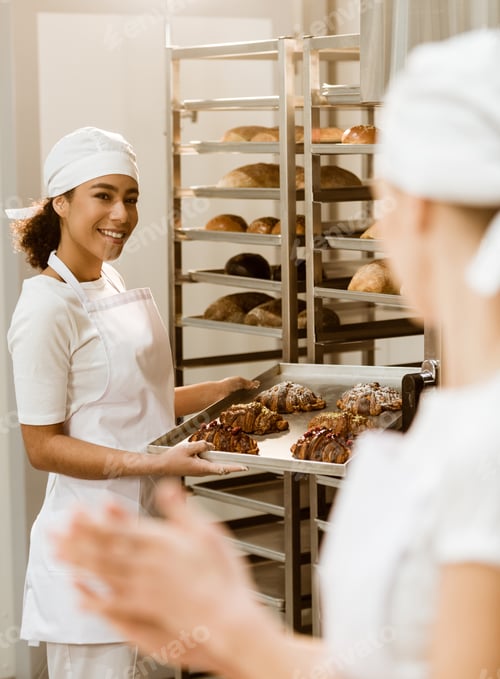 Preview: young female bakers working together at baking manufacture abd talking