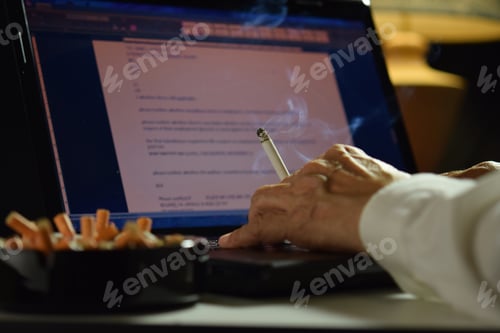 Preview: Man holding lit and smoking cigarette between fingers and working on a laptop computer