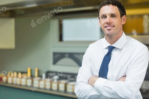 Preview: Mid-adult man standing arms crossed in white shirt, blue tie in kitchen near jars, copy space