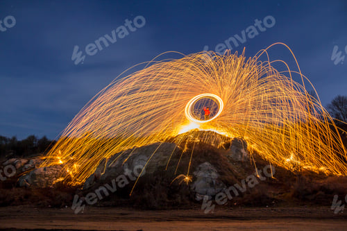 Preview: Long exposure shot of man standing on rocky hill spinning steel wool in circle