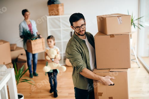 Preview: Mid adult man carrying stack of carton boxes while moving in new home with is family.