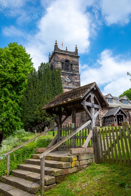 Preview: Historic stone church with wooden lychgate and bell tower amidst lush greenery and bright blue sky