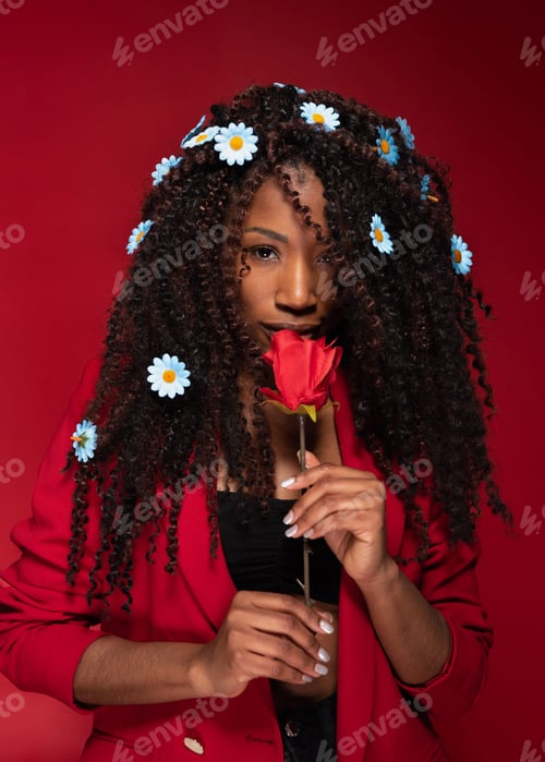 Preview: African american woman with curly long hair smelling red rose with daisies in hair on red background