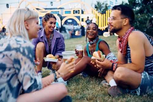 Preview: Multiracial group of happy festival goers eating and drinking beer while relaxing on a grass.