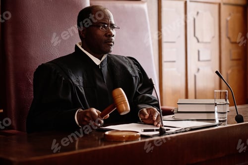 Preview: Experienced male judge with wooden gavel over sounding block sitting by desk