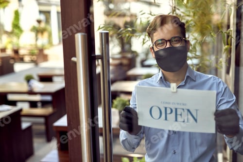 Preview: Happy waiter with protective face mask hanging open sign at the cafe door.