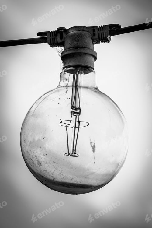 Preview: Grayscale of a light bulb hanging from a thin silver power line cable against the background of sky