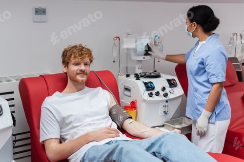 Preview: multiracial nurse in latex gloves and medical mask operating transfusion machine near positive