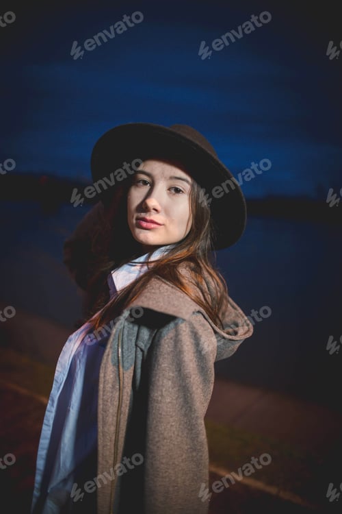 Preview: Vertical shot of a Caucasian female with a hat posing by a river at night