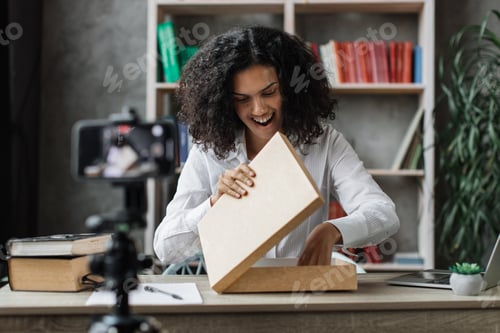 Preview: Pretty multinational woman in casual wear recording video on camera while unpacking gift box.