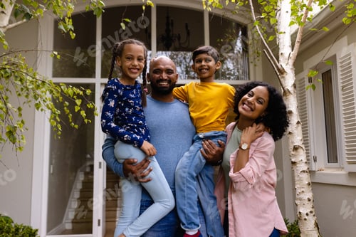 Preview: Portrait of happy biracial parents holding son and daughter, embracing in garden outside house