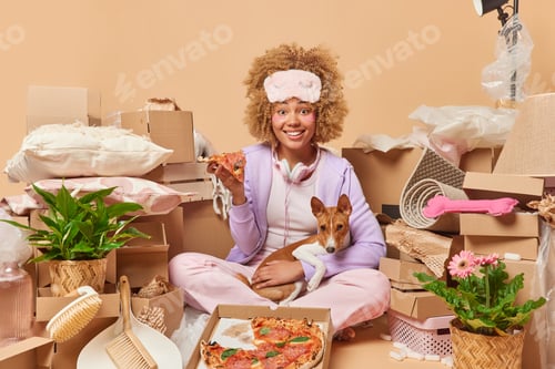 Preview: Positive woman eats tasty pizza poses with favorite pet in new place of living applies beauty hydrog