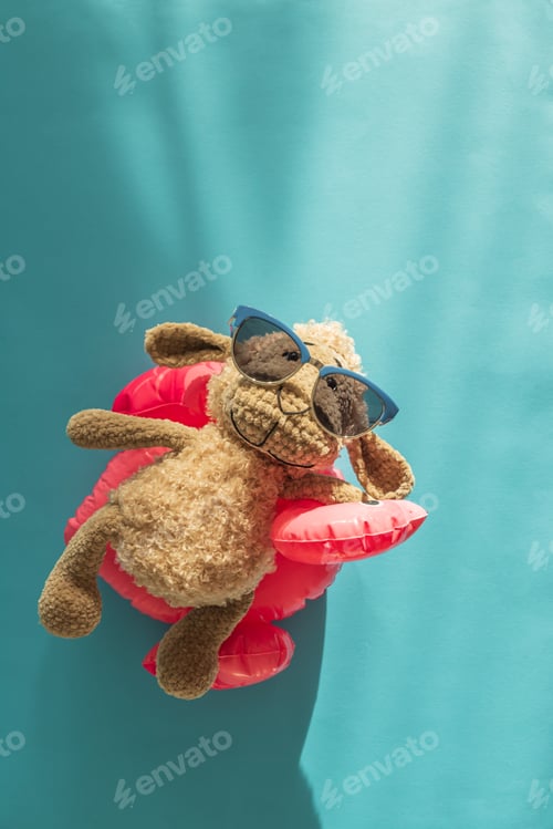 Preview: Knitted Plush Sheep in Sunglasses on Pink Flamingo Float Against Blue Water. Top view