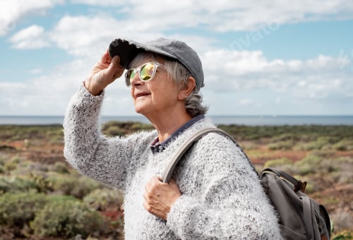 Preview: Portrait of happy senior woman in reflective glasses and hat walking on hiking trip outdoors
