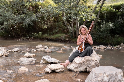 Preview: A young adult woman is playing her indian tanpura sitting on a river rock