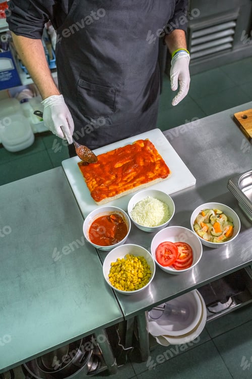 Preview: Anonymous male chef with black apron spreading tomato sauce over focaccia in restaurant kitchen
