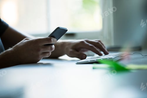 Preview: Hand of male designer using smartphone and typing on keyboard at creative studio desk