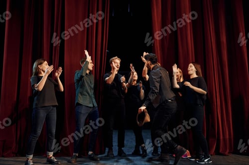 Preview: Standing against red curtains. Group of actors in dark colored clothes on rehearsal in the theater