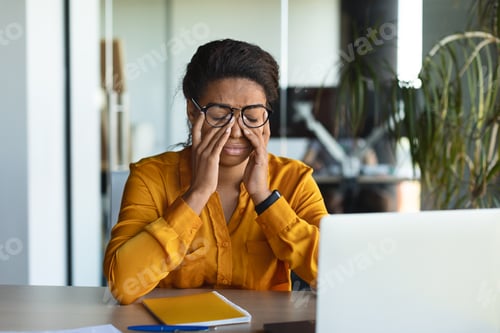 Preview: Tired black businesswoman suffering from headache and fatigue, rubbing eyes at workplace with laptop