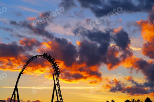 Preview: Black roller coaster is silhouetted against a vibrant orange and pink sunset sky