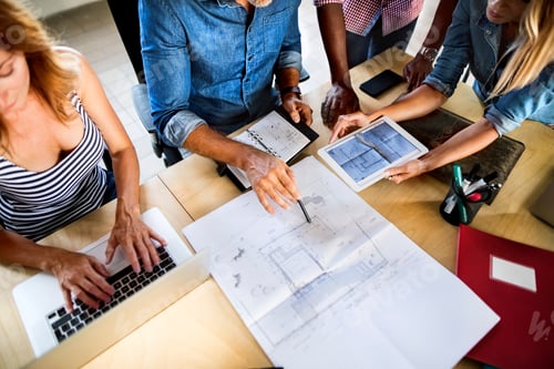 Preview: Colleagues discussing blueprint at desk in office