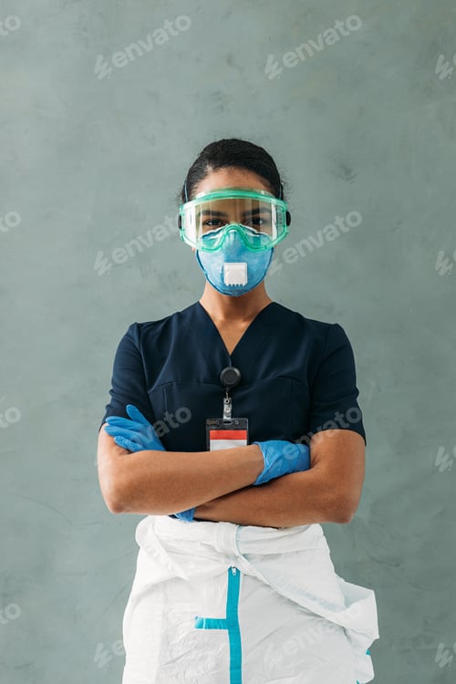 Preview: Young nurse wearing a protective glasses and respirator standing at wall with arms crossed