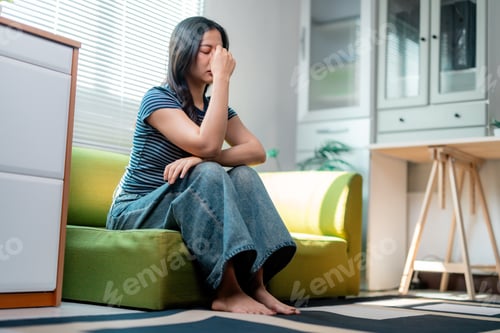 Preview: Stressed young woman suffering from headache, touching her nose bridge while sitting on sofa at home
