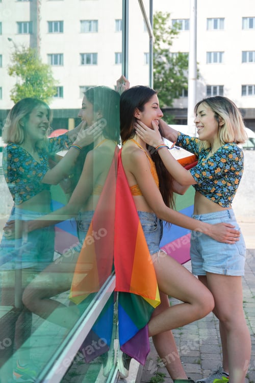 Preview: Young lesbian couple smiling, looking to each other wrapped in a rainbow flag.