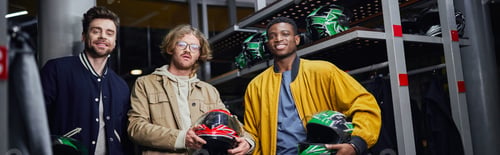 Preview: happy multicultural friends holding helmets and looking at camera inside of racing track, banner
