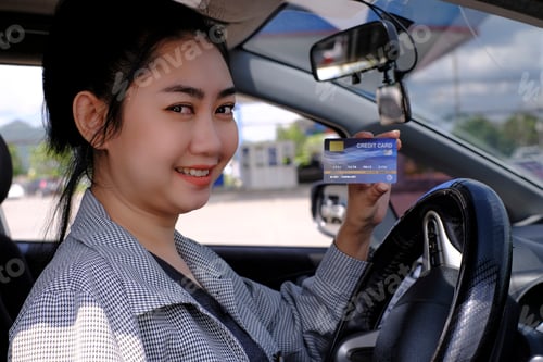 Preview: Happy beautiful Asian woman sitting inside her car showing credit card payment at a gas station