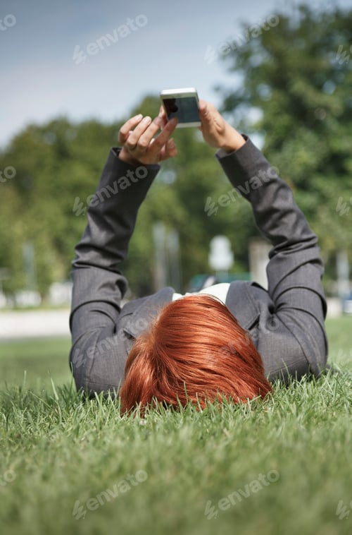 Preview: Surface view of mid adult red haired woman lying on grass using smartphone
