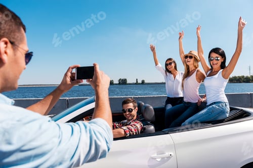 Preview: Capturing happy moments. Rear view of young man photographing his friends sitting in convertible
